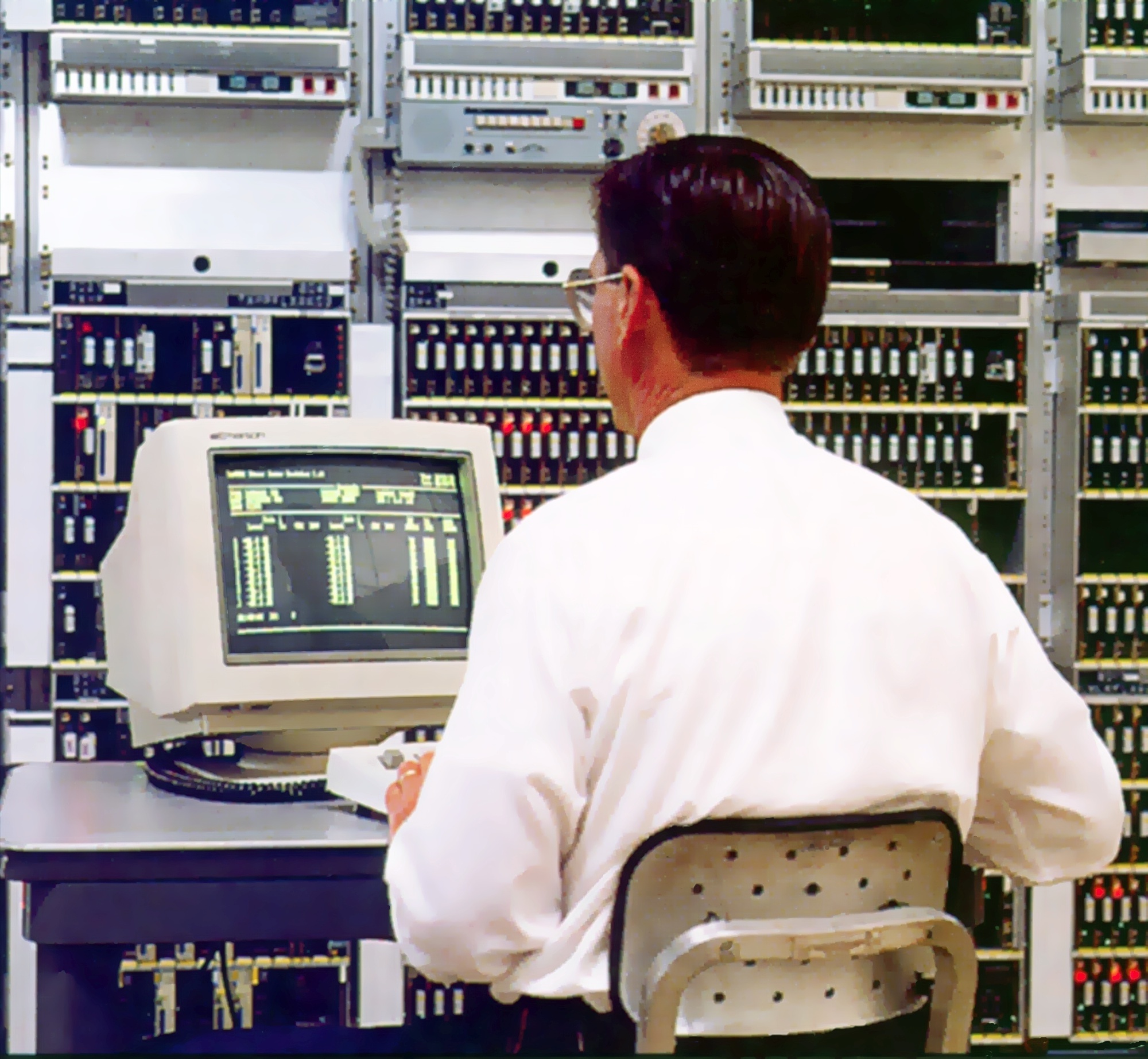 Man sitting at a computer screen with a background of telecom equipment.
